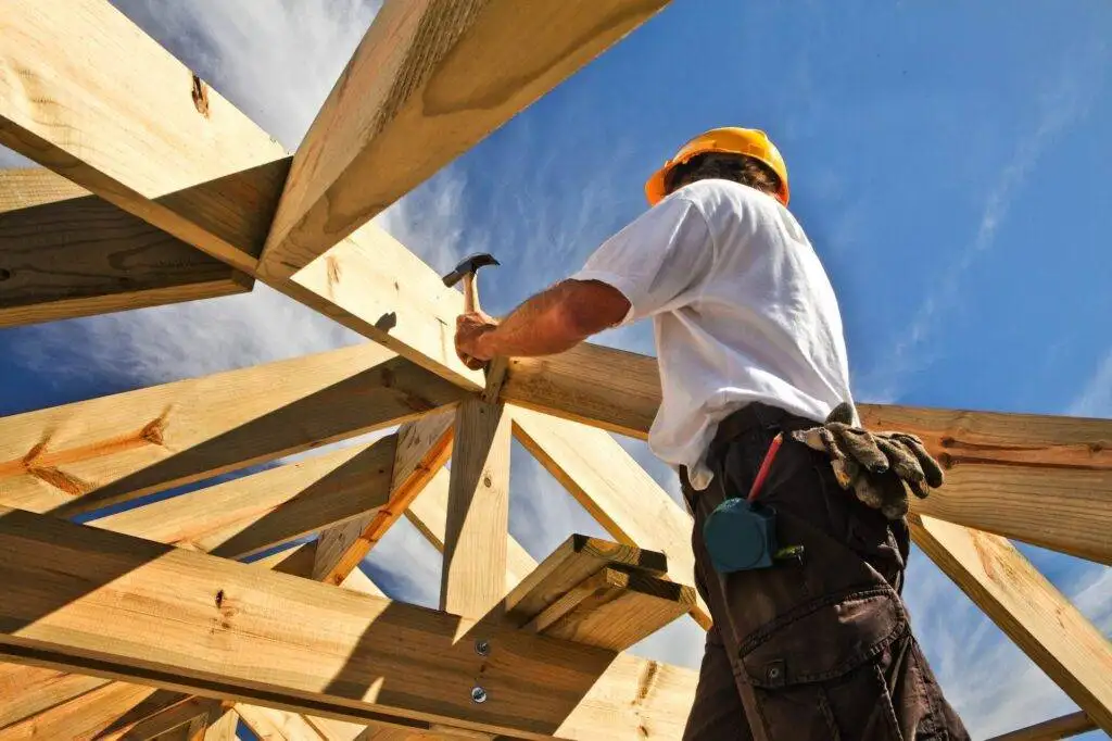 Construction worker in a yellow hard hat hammering a nail into wooden framework